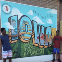 Alum Stands In Front Of Sign That Reads Greetings From Iowa With Another Young Person