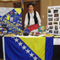 Davor Standing Next To A Poster About Bosnina And Herzegovina Along Witha Table Decorated With The Country Flag And Items From The Country