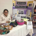 Mira Sitting Behind A Table With A Poster That Reads 22 Musana Orphanage22 And Items On The Table