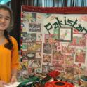 Yes Student Standing By Her Post With Pictures From Pakistan And With Food And Items From Pakistan On The Table