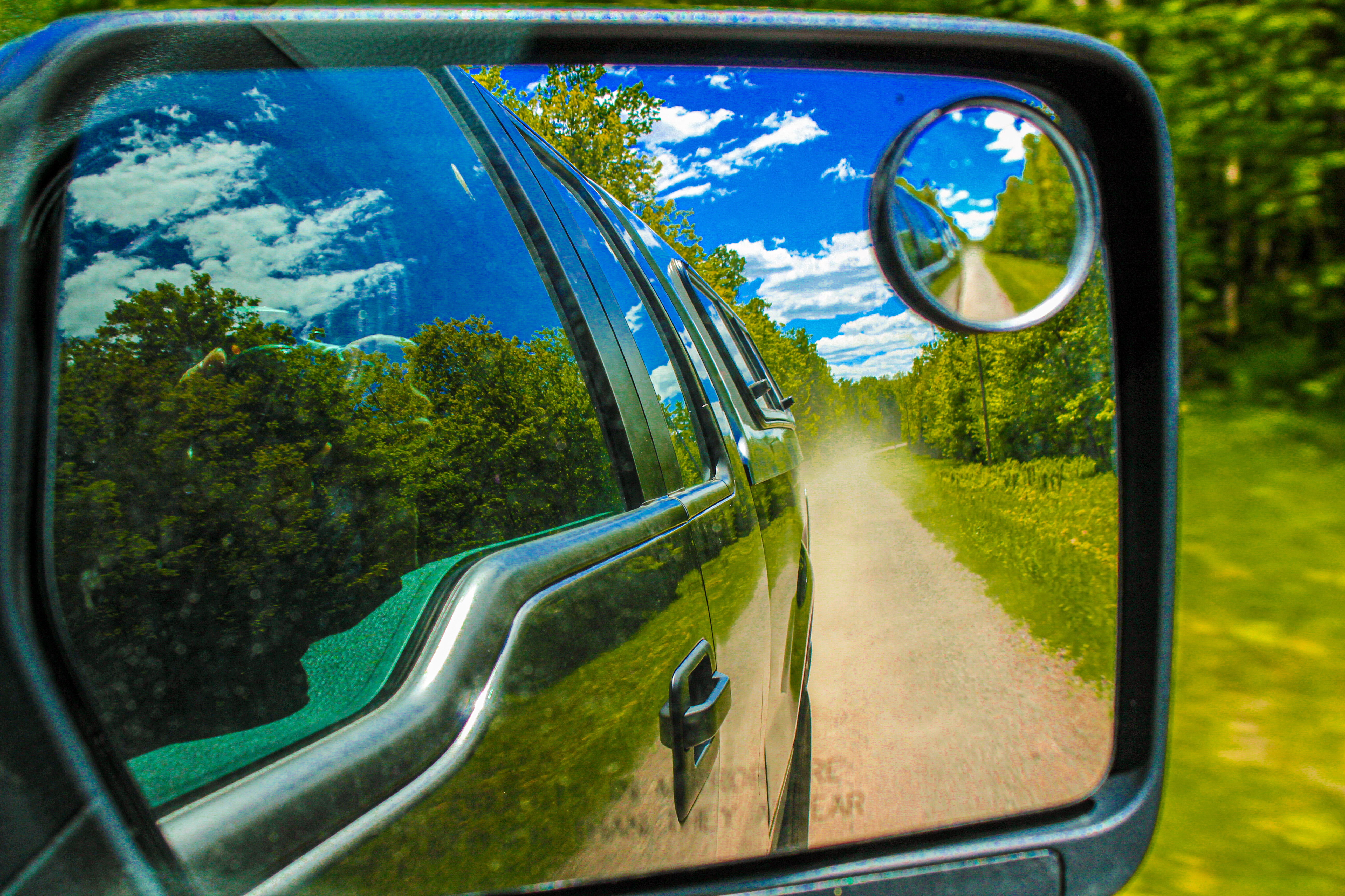 Photo of a sideview mirror of a car reflecting a dirt road, trees, and blue skies