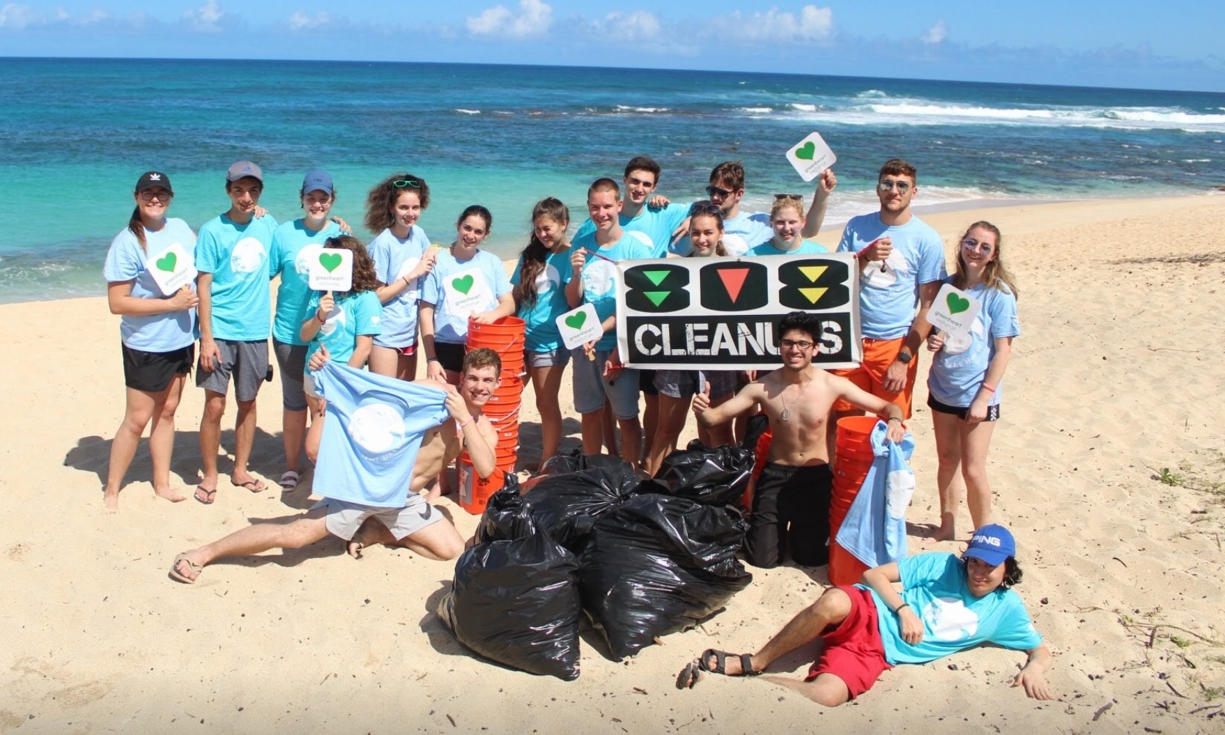 A large group of students stand on the beach in front of bright blue water with bags of trash.