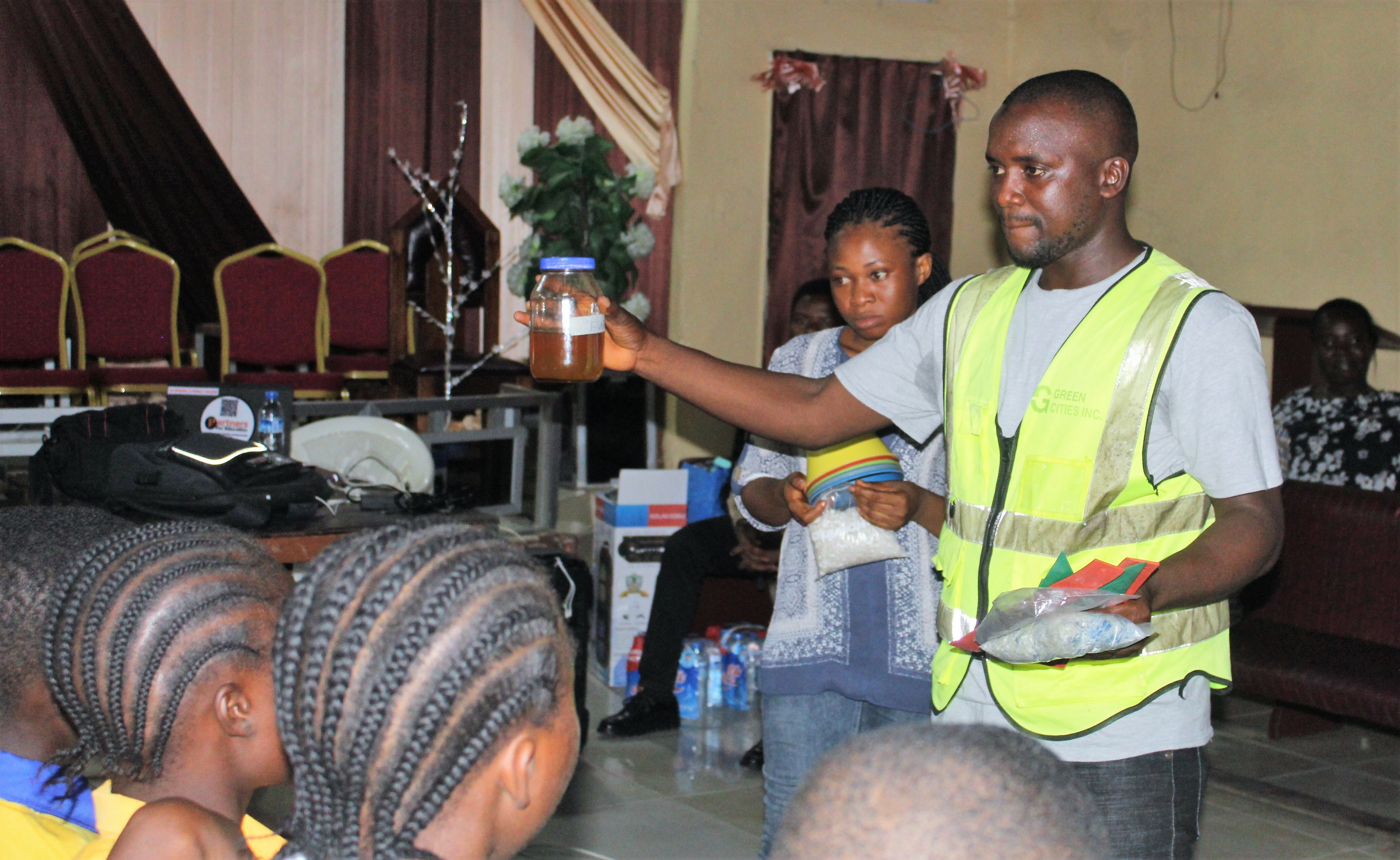 A Facilitator Is Standing In Front Of The Room Presenting And Displaying A Product That Can Be Made From Waste