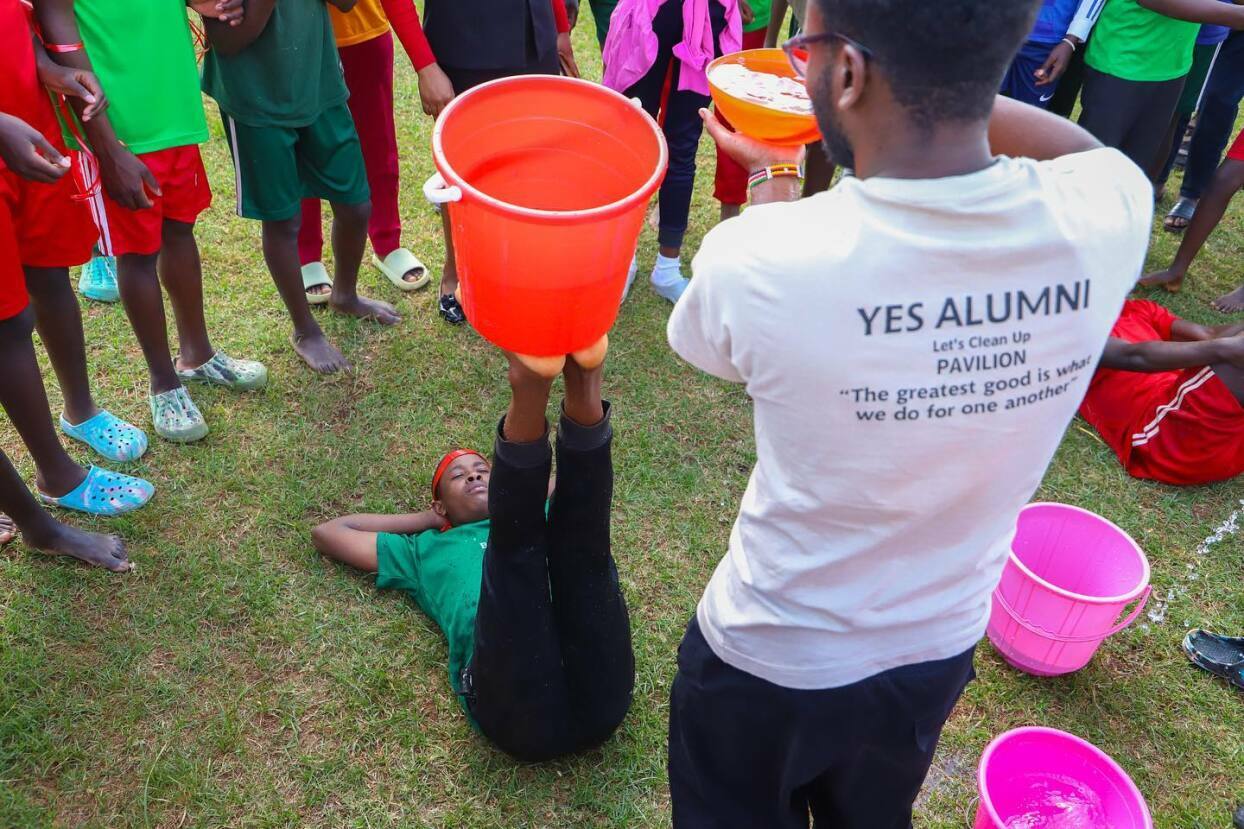 A male guide with YES Alumni writing on the back pouring water in a bucket while one of the male participants is holding the bucket with his feet