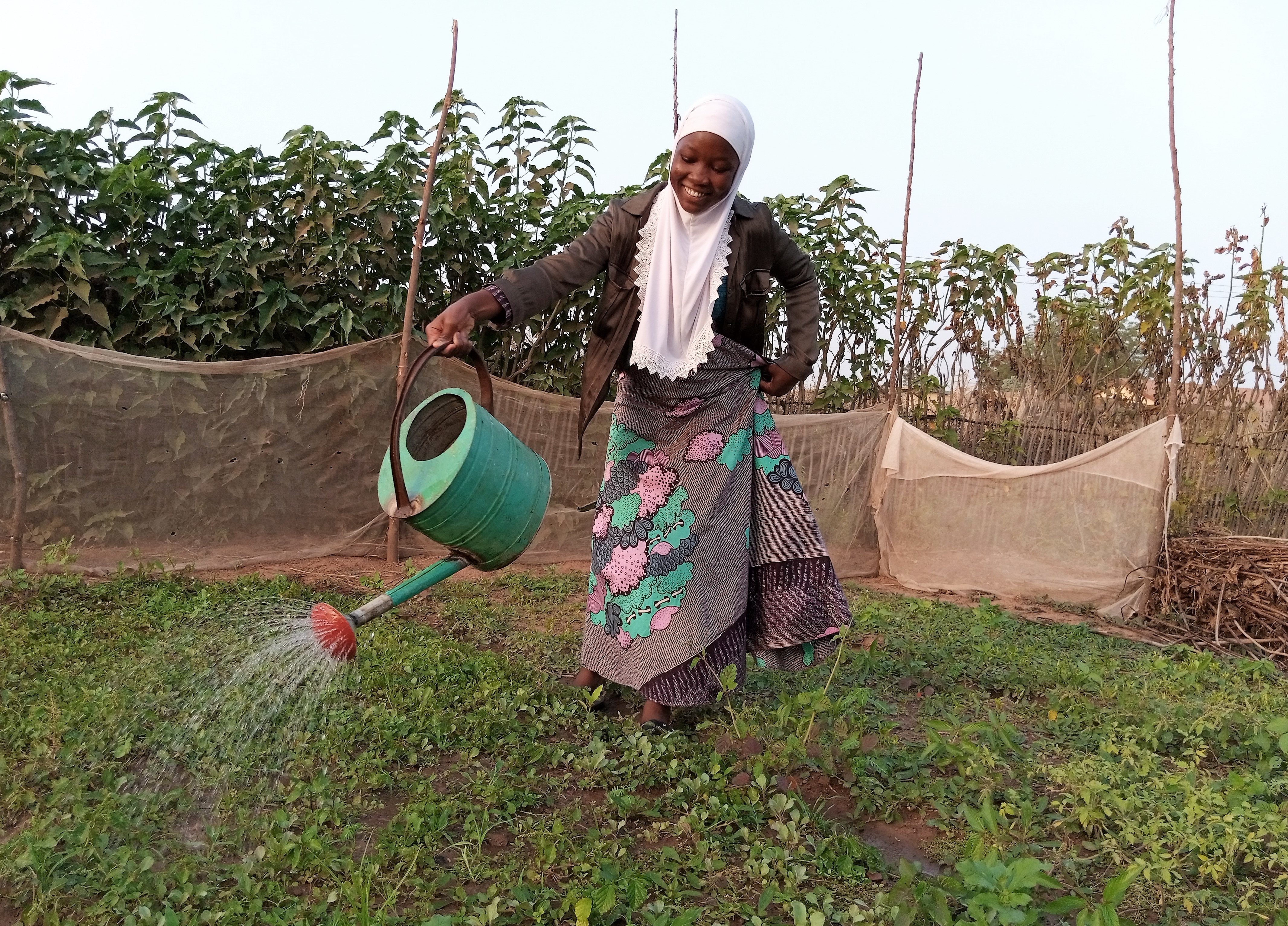 Inusah smiling, holding a watering can and watering a vegetable nursery