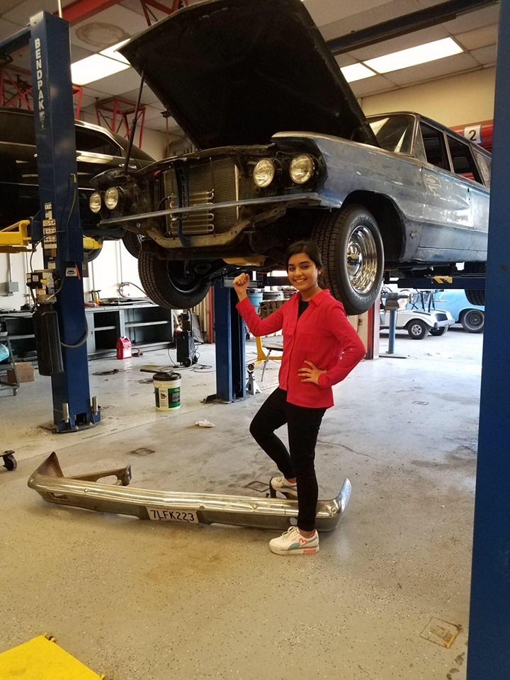 Amna stands underneath a car that is propped up for automotive construction class
