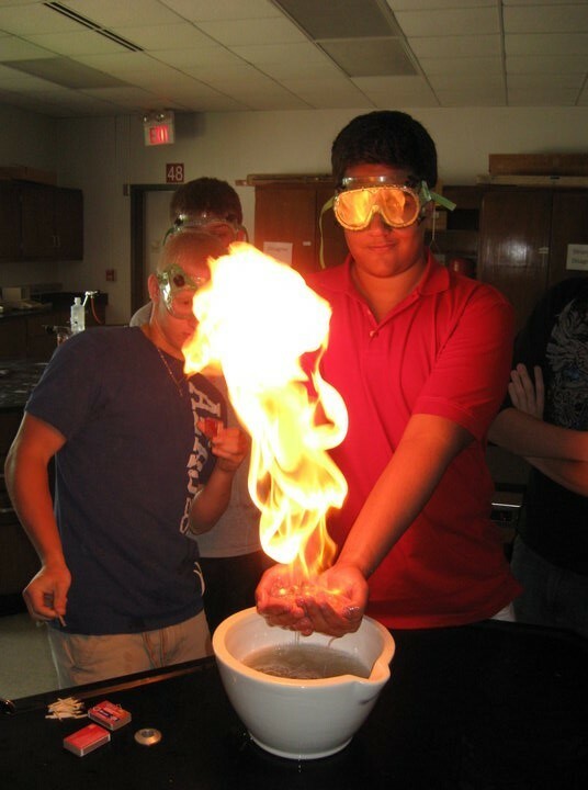 Abdulaziz and another student stand with goggles on with a large fire sparked in front of them