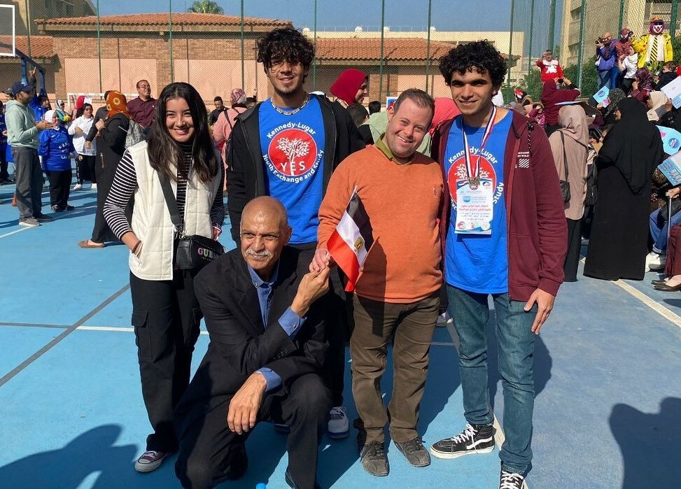 Five people, including two alumni in YES shirts, pose for the camera with many community members enjoying festivities in the background.
