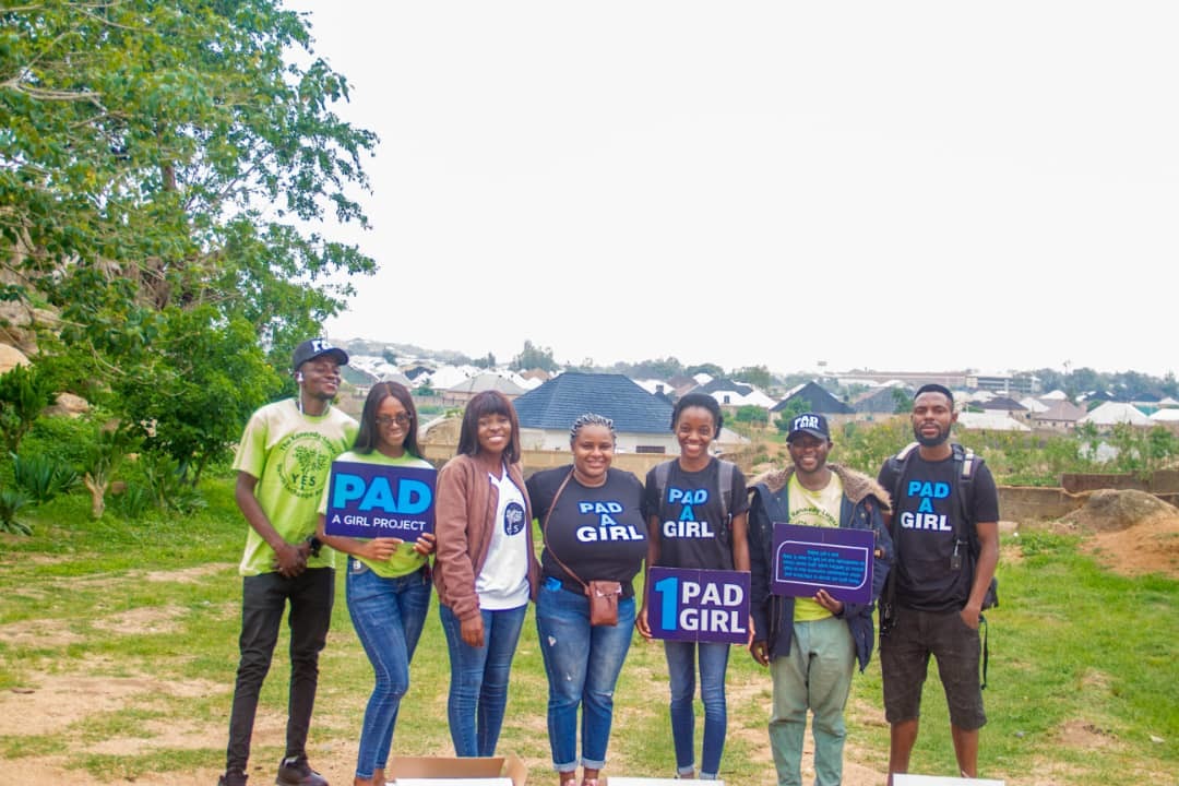 Eight people stand outside with signs that say "PAD GIRL"