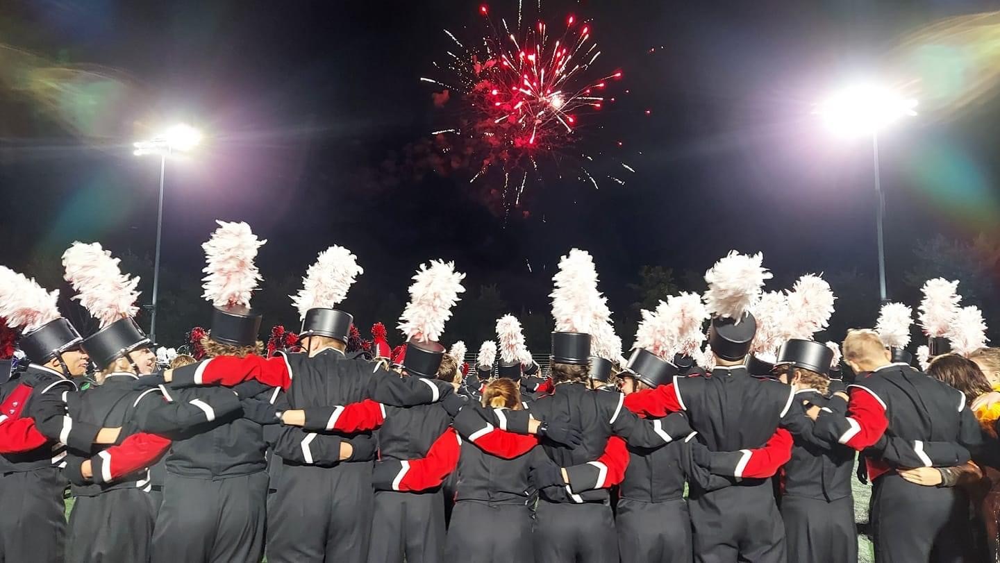 Dozens of teens in black and red band uniforms link arms and watch fireworks