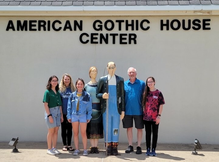 Four teen girls and an adult man stand with statues of farmers in front of a white building labeled American Gothic House