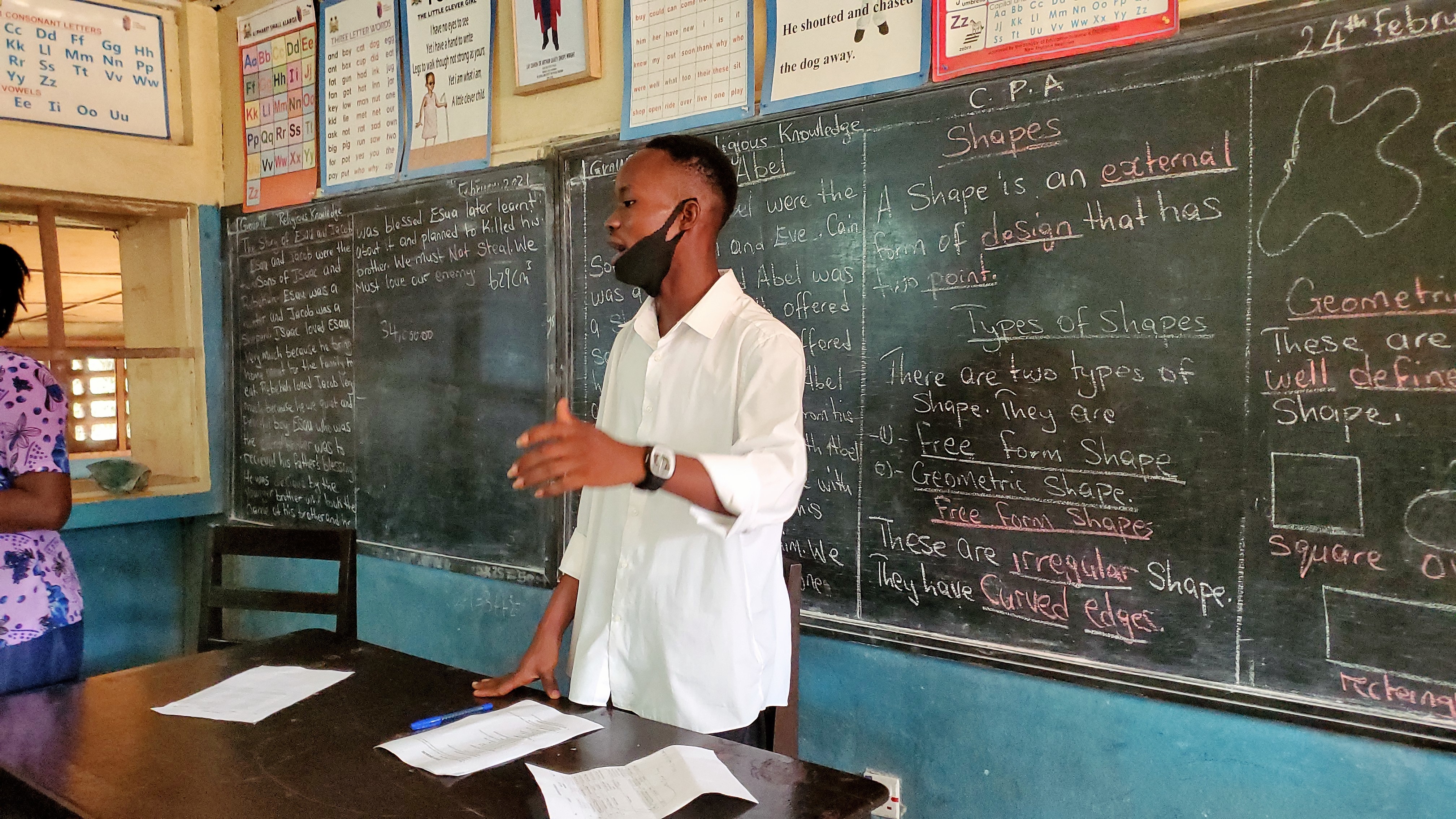 YES alumnus Koroma stands in front of a classroom lecturing. A blackboard full of text is behind him