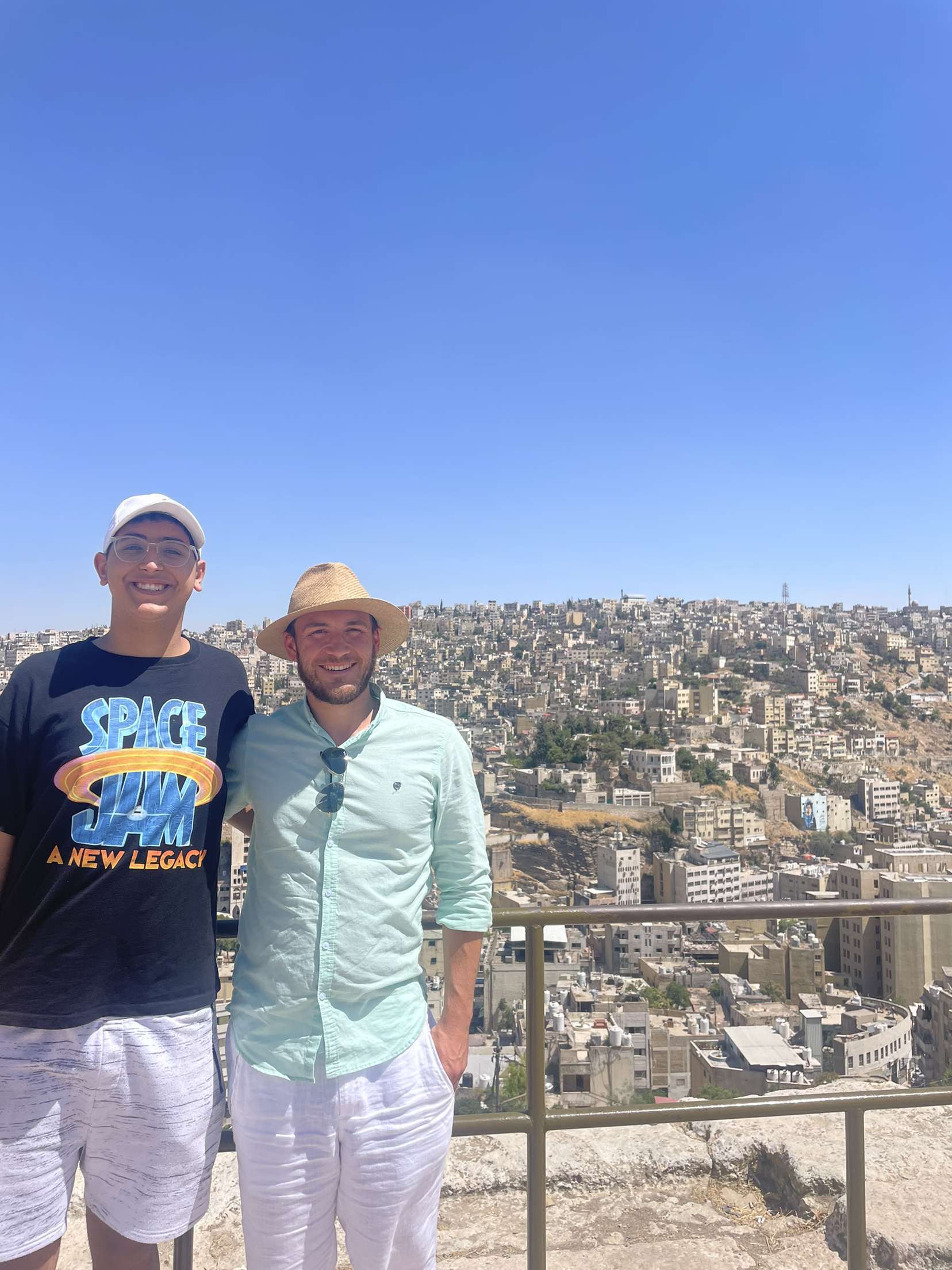 Mohanad and James smile and pose together in front of a blue sky and view of Amman