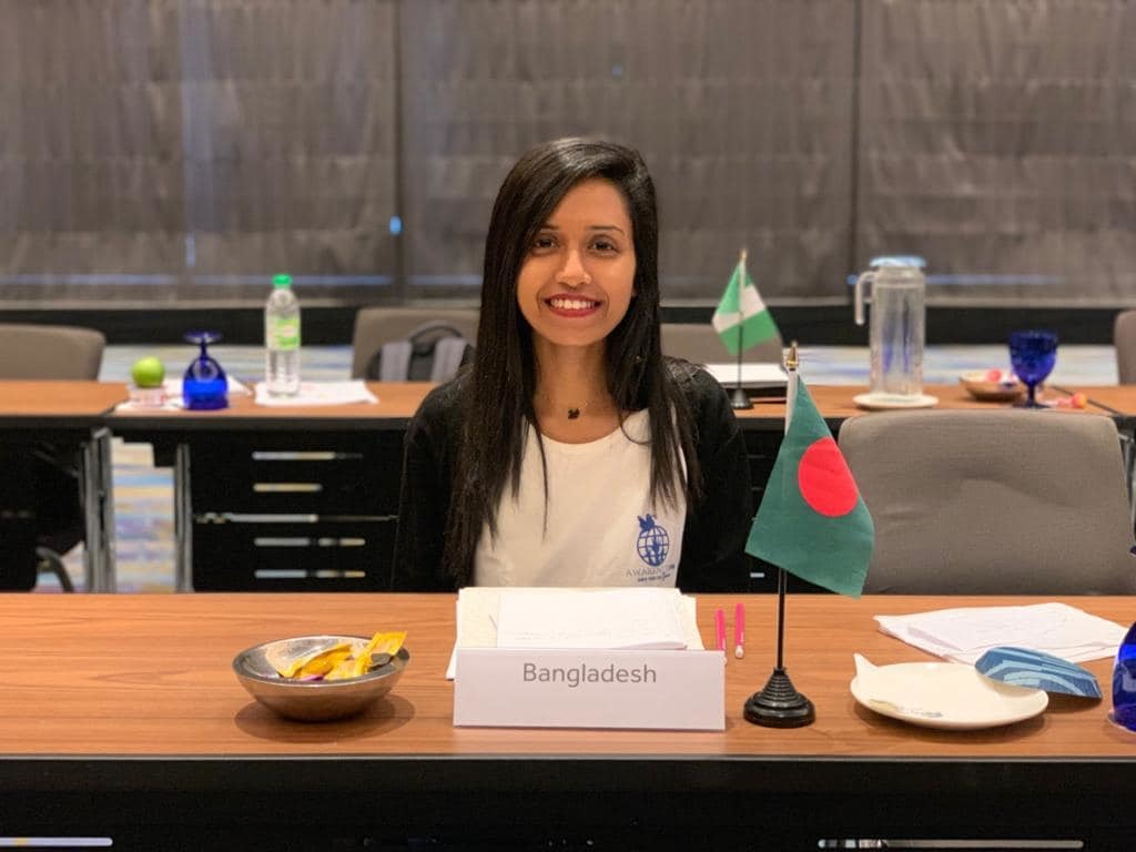 Shomy sitting at the United Nations with a Bangladeshi flag in front of her