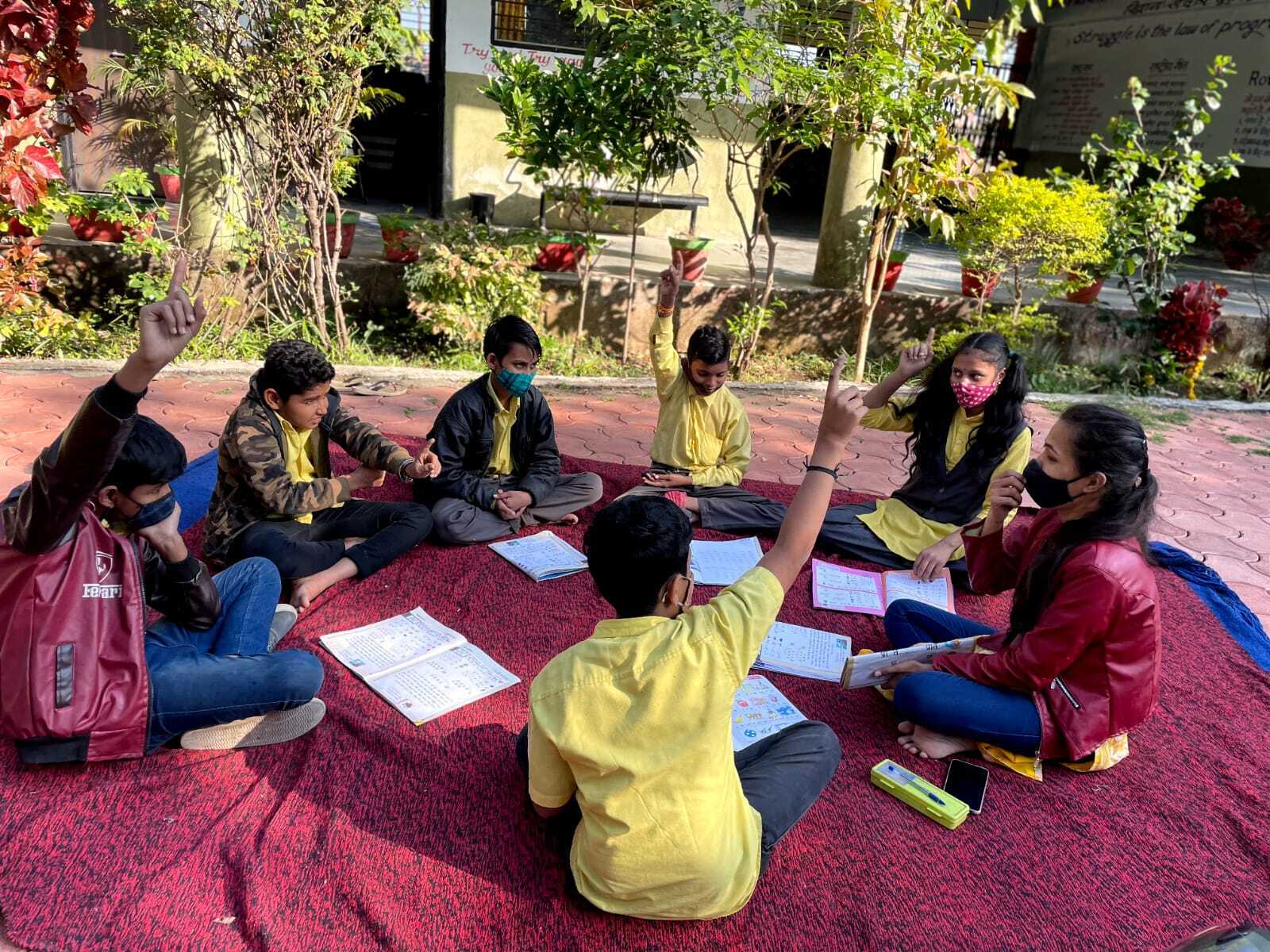 Six Children And An Instructor Are Sitting On The Ground In A Circle Studying  Three Children Are Raising Their Hand To Answer A Question