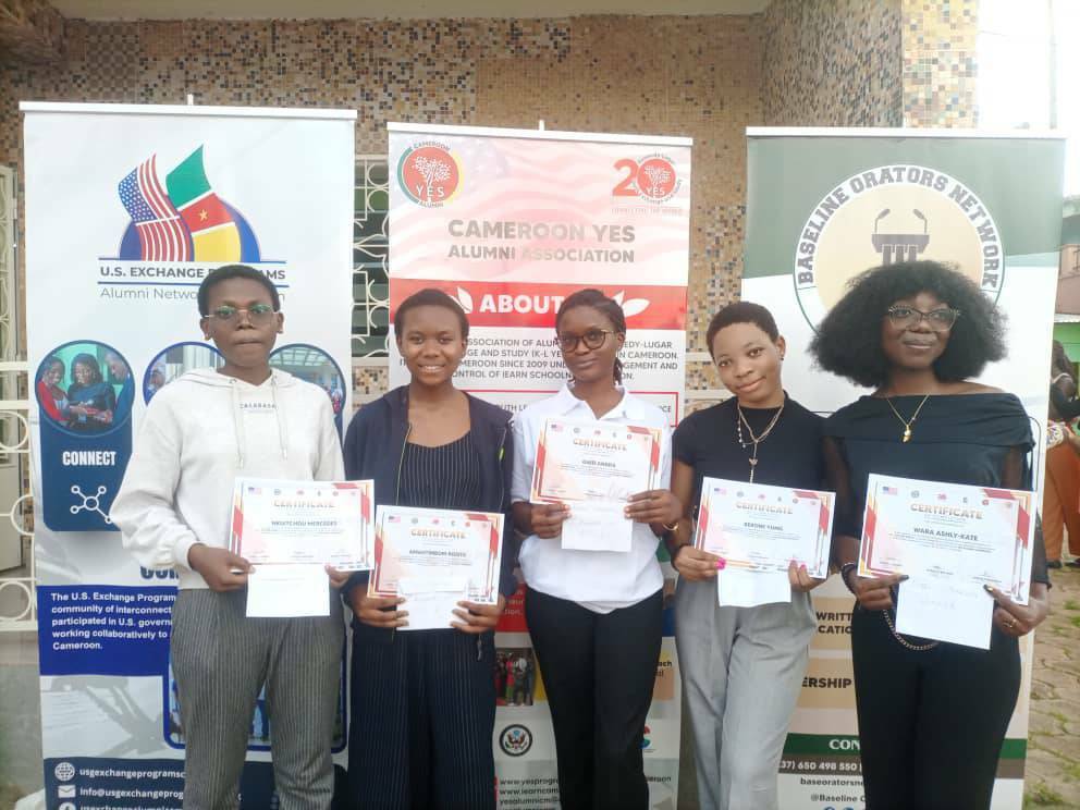 The five winners of the debate and public speaking contest posing in front of banners with certificates and prizes