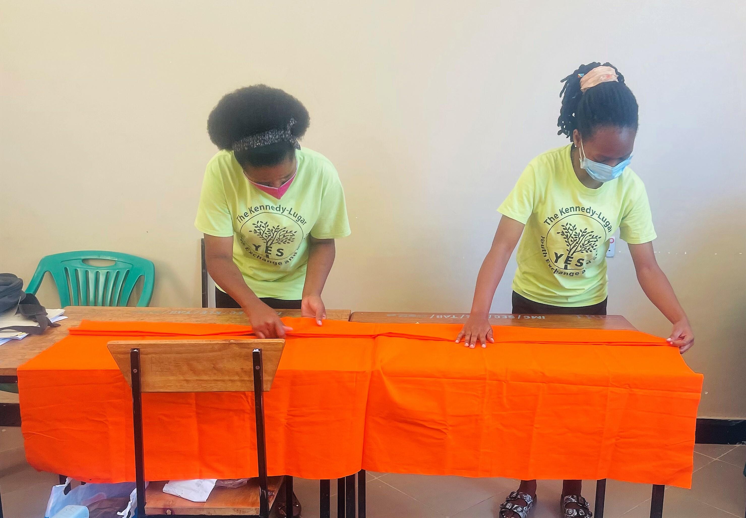 Two Alumni Team Members Are Standing Behind A Table Demonstrating Tie Dye Steps For Making Colorful Patterns Batik On A Piece Of Cloth