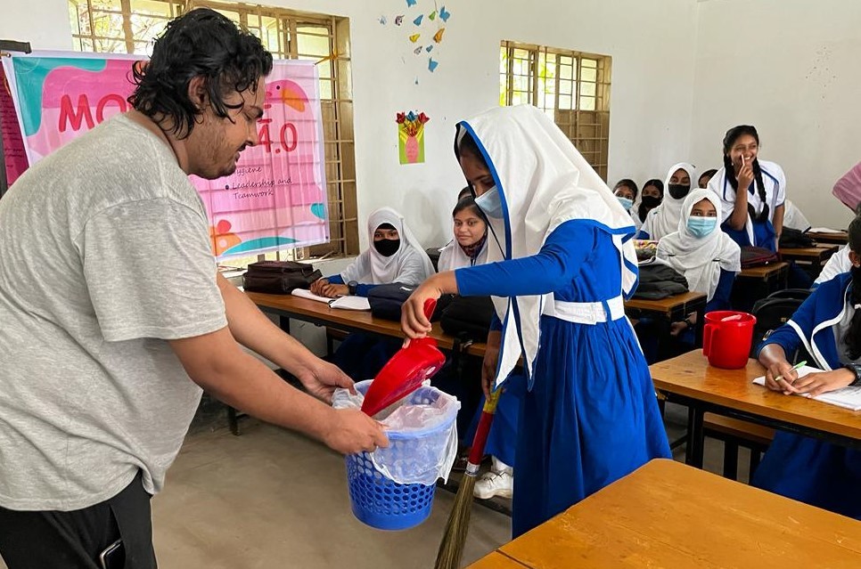 Yes Alumnus From Bangladesh Is In A High School Classroom Holding A Dustbin For A Student As She Empties Dirt Into It