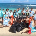 A large group of students stand on the beach in front of bright blue water with bags of trash.