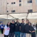 6 Alumni Standing In Front Of A Tan Building And Tent