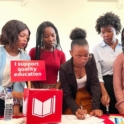 A group of people standing around a table with a sign "I support quality education" filling in paper forms