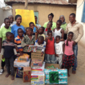 A Group Of Young Children And A Few Adults Standing In A Group Smiling  In Front Of Them Are Bags Of Donations Clothes Diapers Etc