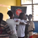 A group of young girls gathered around a woman who is demonstrating a physical movement with a YES program banner in the background
