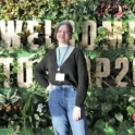 Armela in front of a sign that reads "Welcome to COP26" surrounded by greenery and flowers. 
