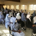 A large group of female students sit in the classroom with masks and hijabs on.