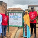 Five Alumni Project Team Members Pose For A Photo At The Project Sign By The Well