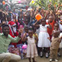 YES alumnus, Joseph, is kneeling at the front of a group of children who are holding up their hands and waving balloons.