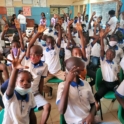 Inside a classroom a group of students in uniforms raise their hands.