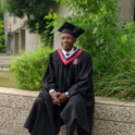 YES alumnus, Amisse, sitting on a ledge outside in a graduation cap and gown. 