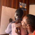 One person writes on a paper taped to anothers back with the front person drawing on a wall mounted sheet while the second writes on a separate paper