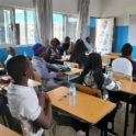 Photo Shot From Back Of Classroom Where A Group Of Students Face Forward To A Project Volunteer Speaking In Front