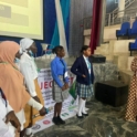 Project leader holding a microphone and standing with participants in front of the stage with a slide asking what they have learned