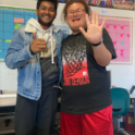 Roepesh standing with a friend in a classroom, waving to the camera.