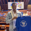 YES alumnus, Mamadou holding a blue YES T shirt at the World Forum for Democracy.