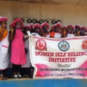 A large group of women hold a banner. They wear bright pink and white aprons and hats.