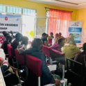 Facilitator writing on a flip chart while participants, including a woman holding a baby, are seated and listening attentively