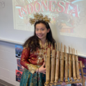 Yvonne smiling in front of a presentation about Indonesia, dressed in traditional clothes and holding an instrument