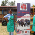 Two female alumni stand on either side of a YES alumni banner that says Thank you YES