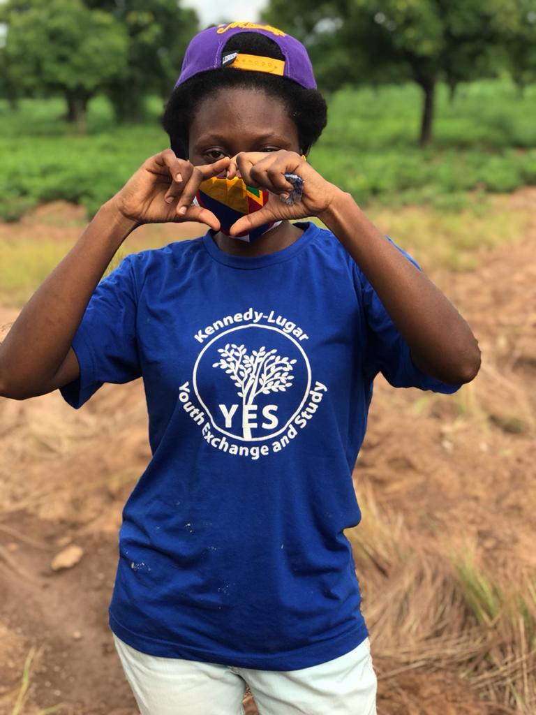 A participant wearing a blue YES shirt stands in a field making a heart with her hands.