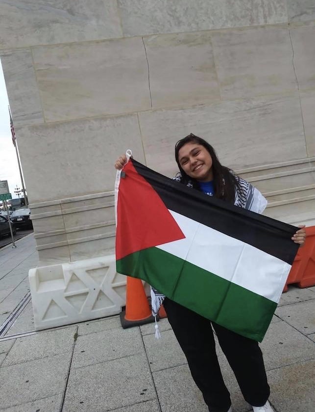 YES student, Saba, standing in front of a concrete building holding up the Palestinian flag.